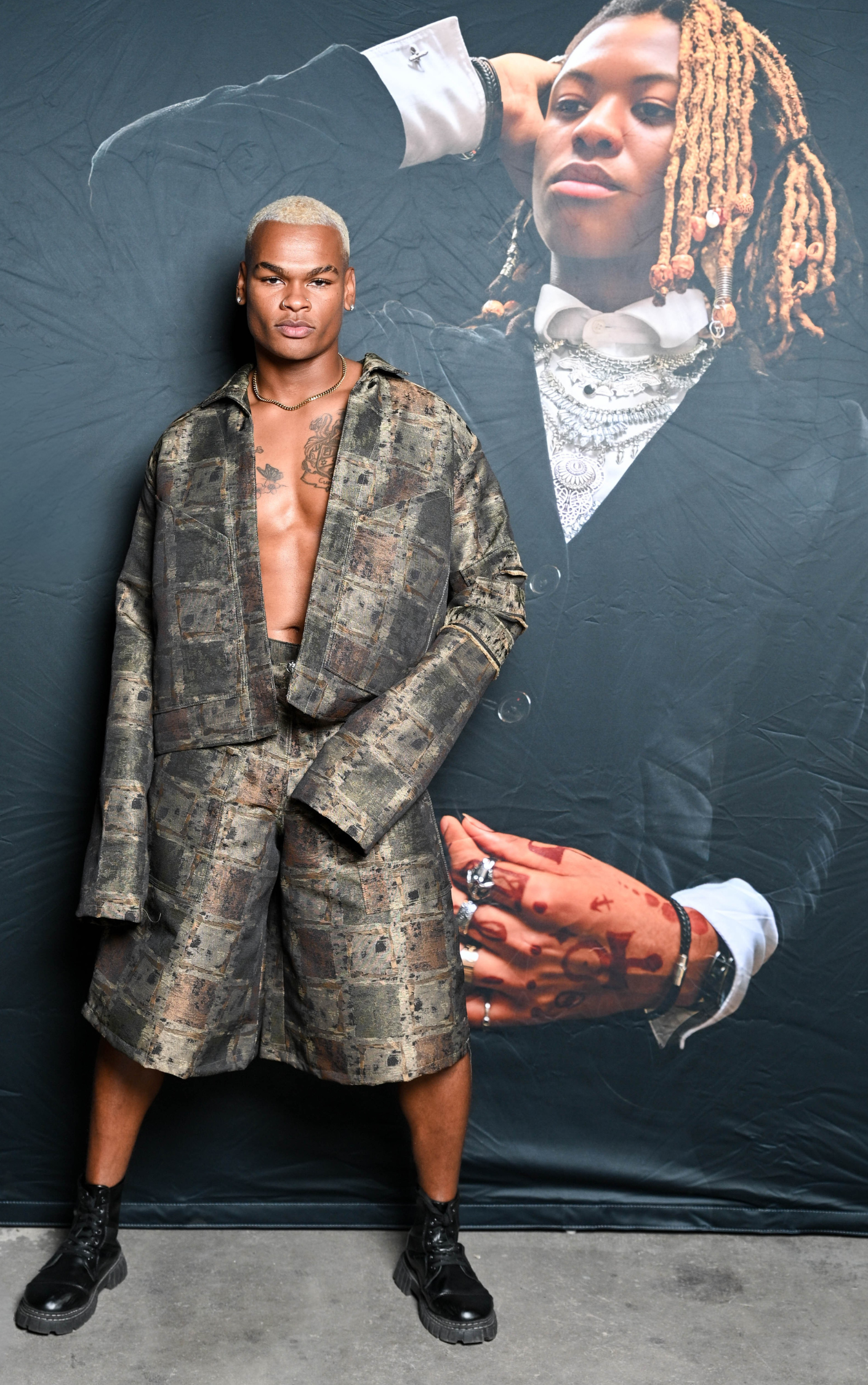 Model wearing 14th Pharaoh R.A.W. Painter Bark Jacket and Jorts camouflage outfit standing in front of a large image of Matthew E. Bell, designer in formal attire.